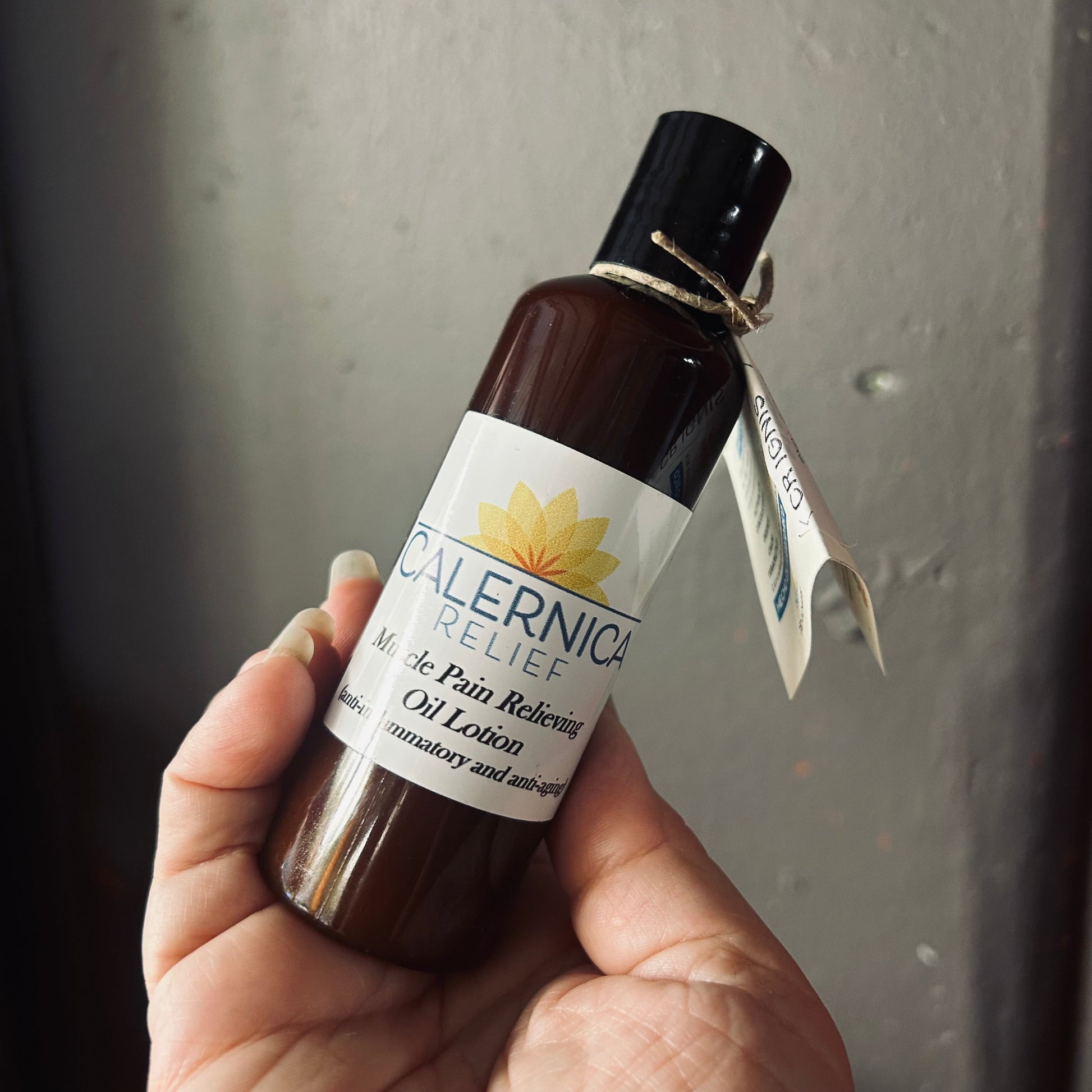 Hand holding a bottle of Calernica Relief Muscle Pain Relieving Oil Lotion against a gray background